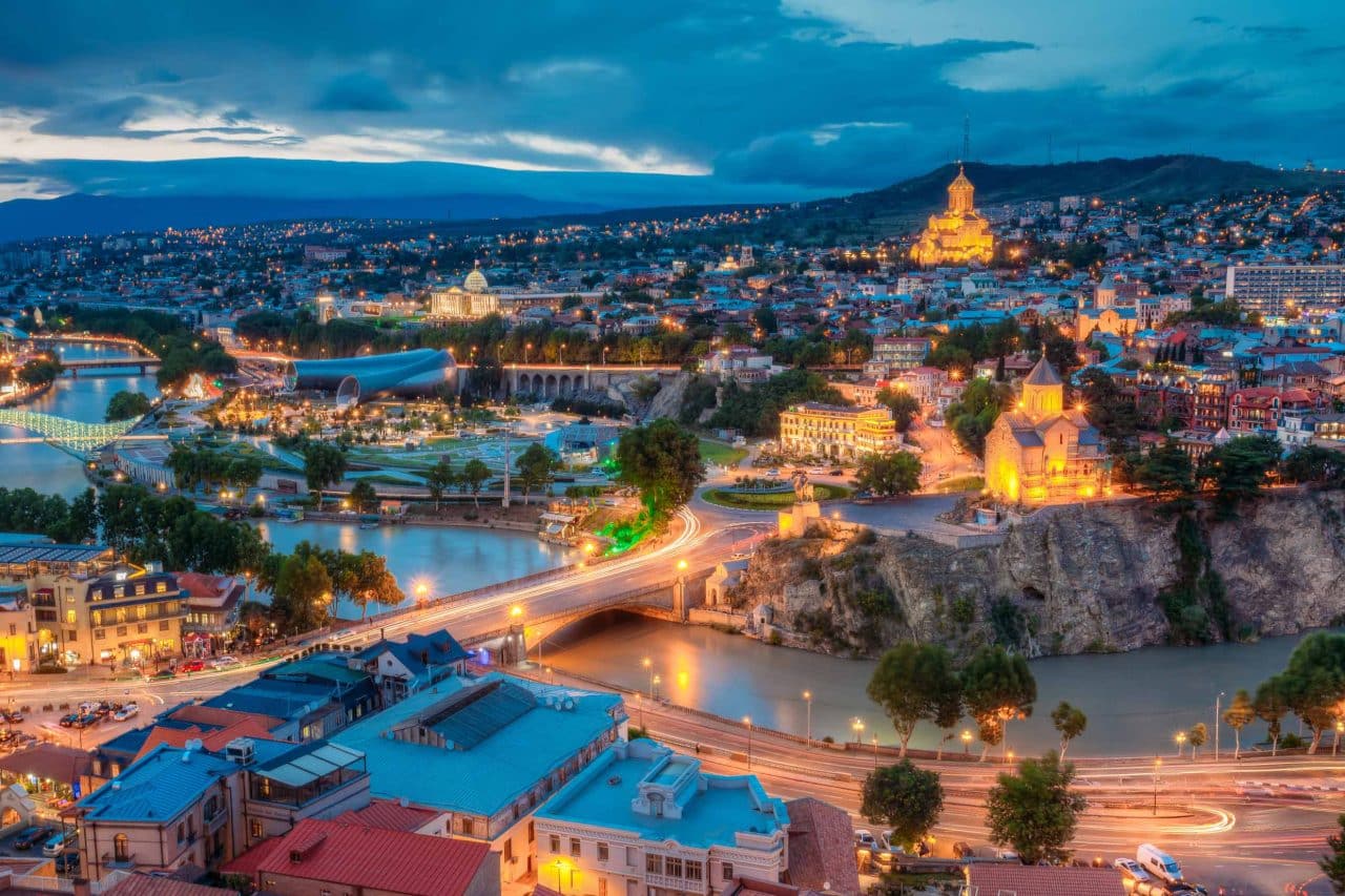 Scenic Top View Of Central Part Of Evening Tbilisi Georgia With Scenic Top View Of Central Part Of Summer Evening Tbilisi, Georgia. Metekhi Bridge And All Famous Landmarks, Sightseeings In Illumination Lights. Dramatic Blue Cloudy Sky.